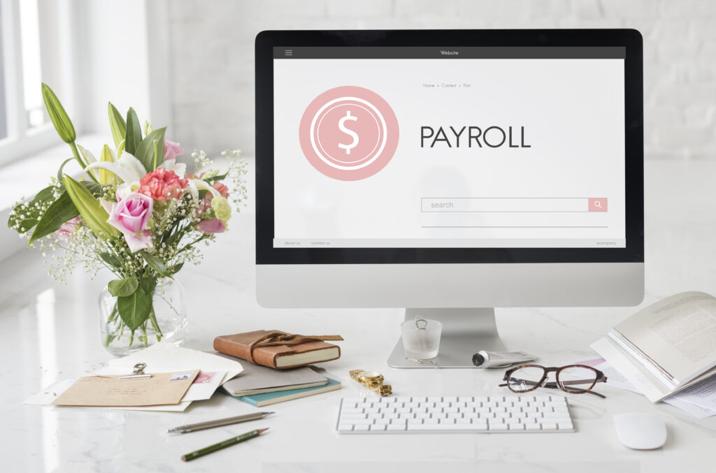 Computer screen displaying payroll concept with dollar icon, surrounded by office desk items like flowers, notebook, and glasses in a bright workspace.
